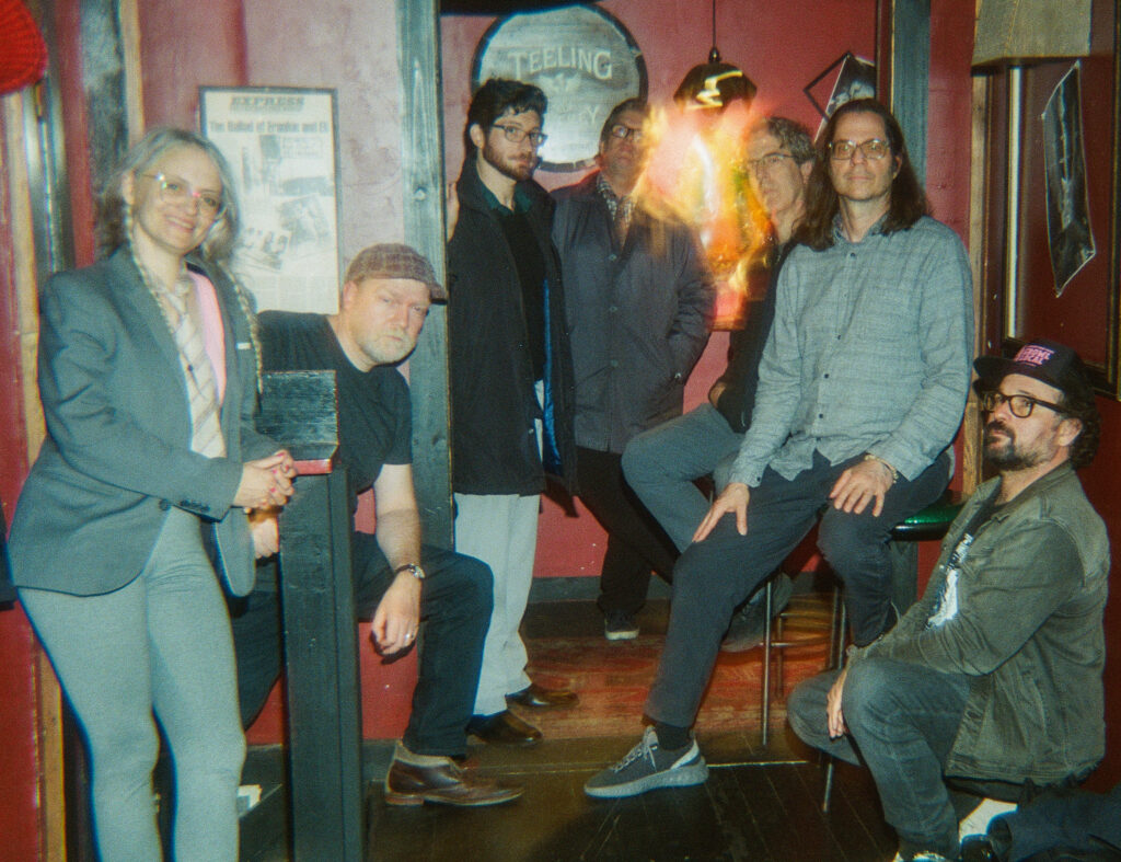 A photo of Inner Ear Brigade's 7 2024-25 cast members, taken in the dark but cozy and expansive Eli's Night Club green room. The photo is taken with an analog film camera, and there are some pleasant warm light streaks around a lamp's reflection in a mirror in the back of the shot that illuminates the red paint of the walls.