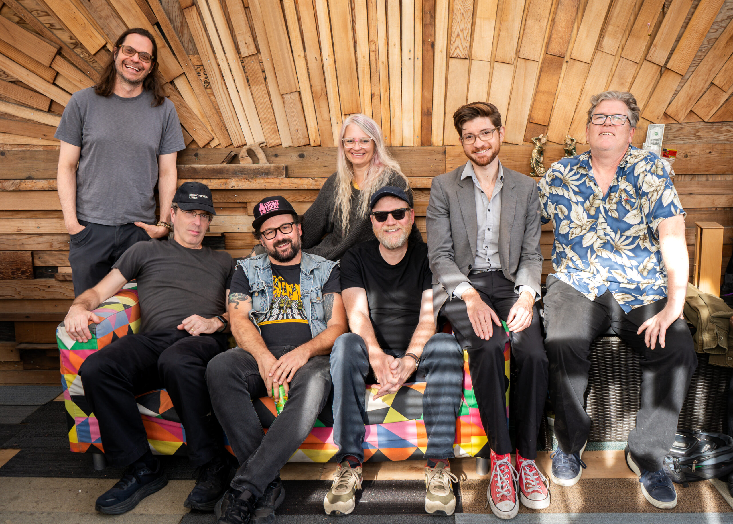 Photo of 7 people in Inner Ear Brigade's 2024-25 lineup, smiling at the camera. They are all arranged on, by and on top of a couch with beautiful wooden inlay radiating out from the center behind the group.